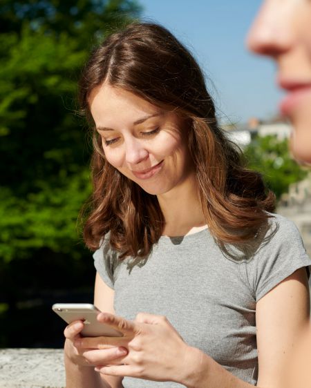 Junge Frau blickt auf Handy, im Vordergrund weitere Frau unscharf zu erkennen