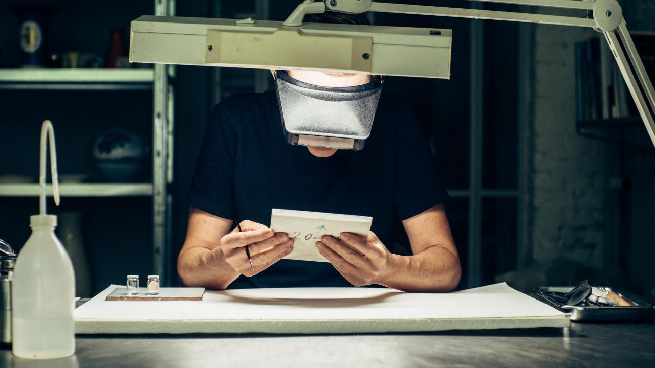 A restorer with protective glasses sits at a workbench, examining a white ceramic object under a desk lamp