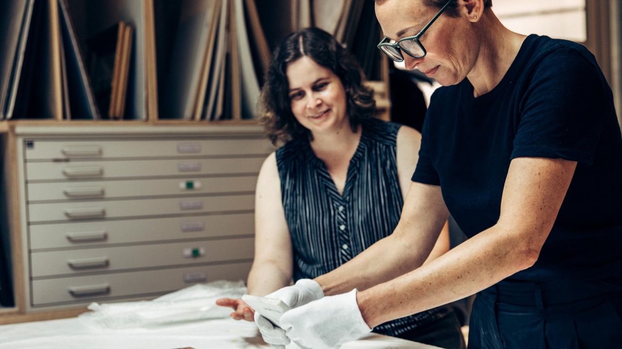 A restorer wearing white gloves stands at a work table holding a piece of porcelain in her hands, while a second woman watches with a smile.
