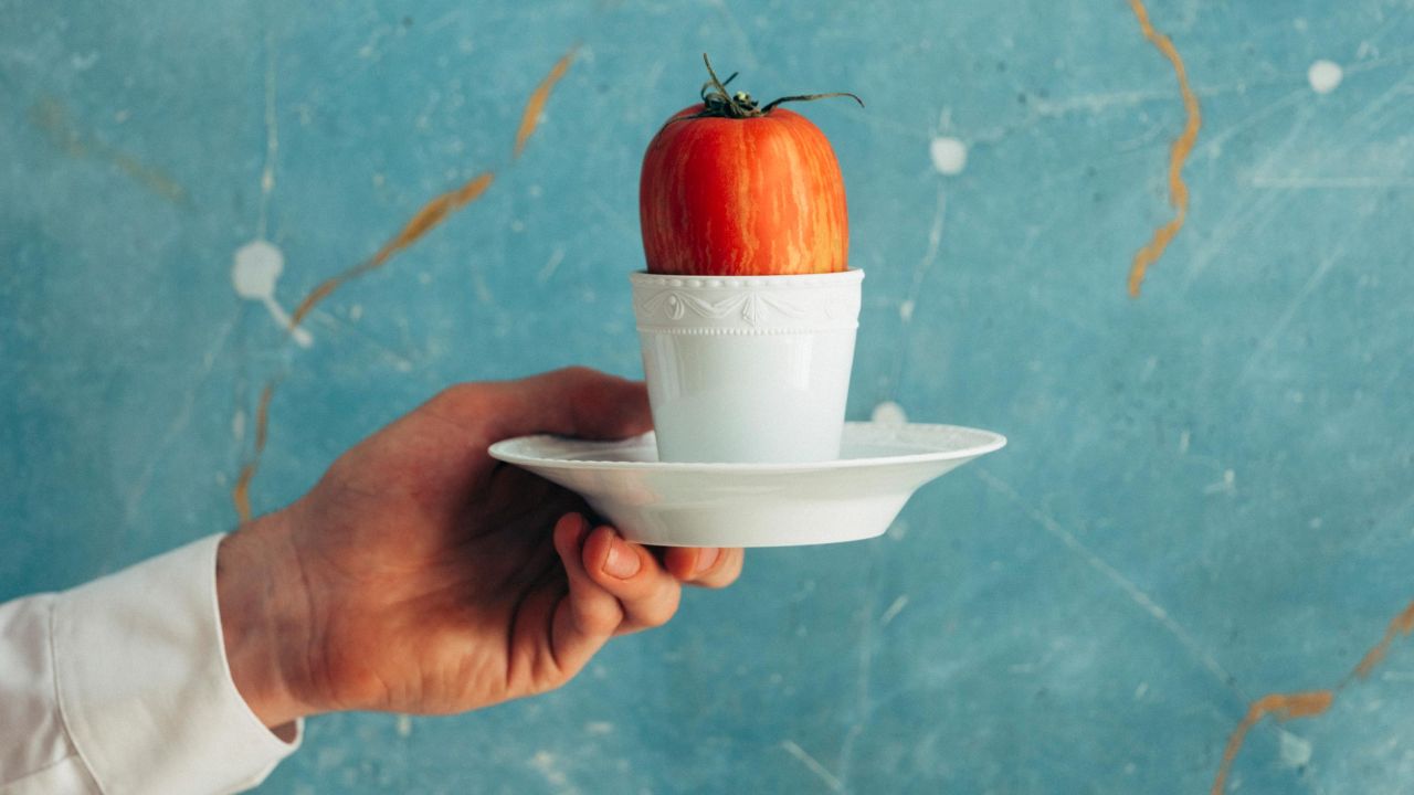 A person is holding a cup with a tomato in their hand, with a blue and brown marble wall in the background.