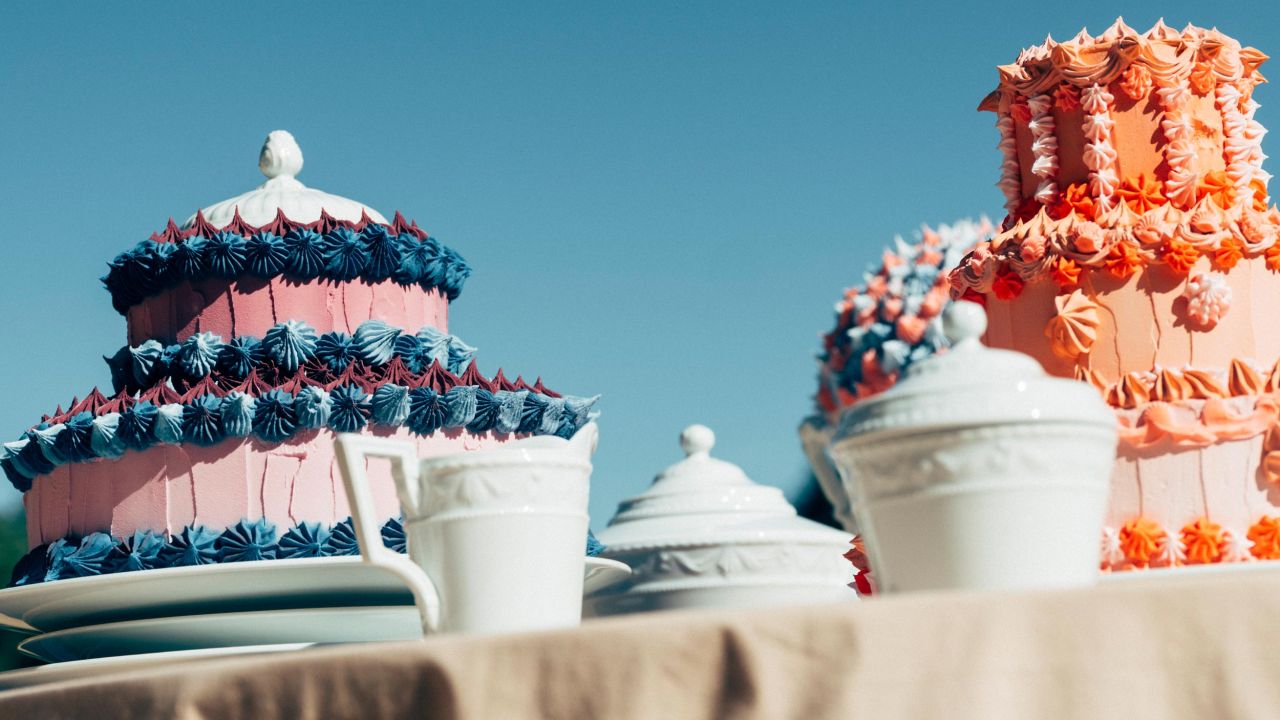 Two large, elaborately decorated cakes and KPM porcelain on a table outdoors under a blue sky