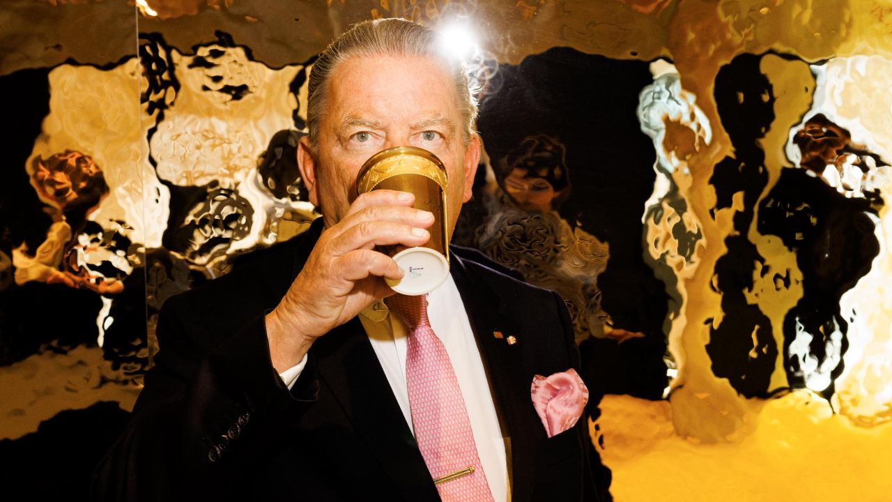 Close-up of a man in a suit and tie drinking from a golden cup, with a highly reflective golden wall in the background