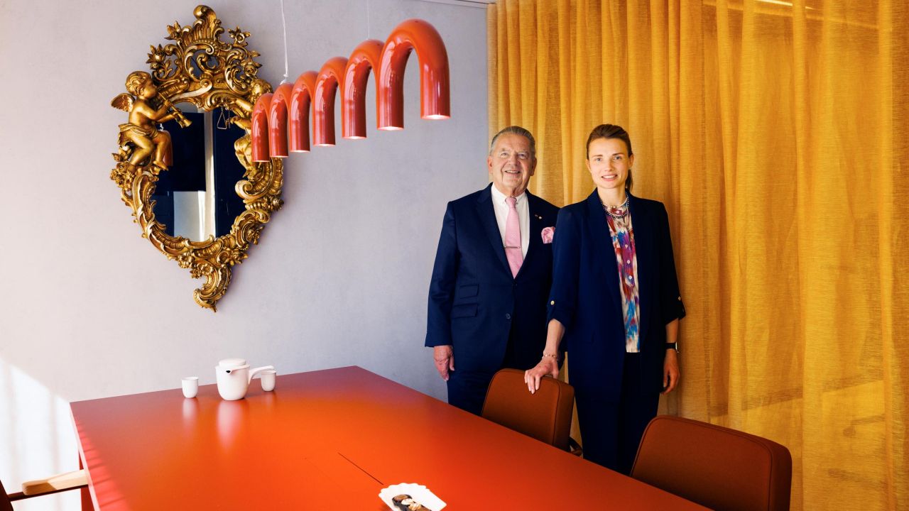 A smiling man and woman pose in a modern conference room in front of an orange table and yellow curtains
