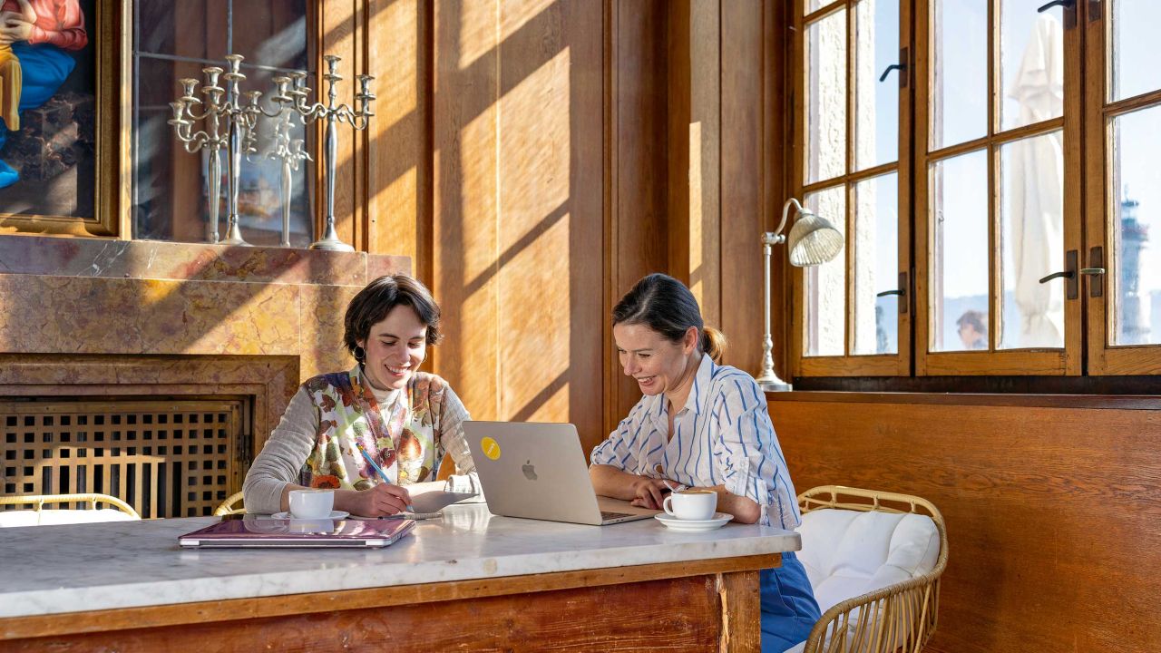 Zwei Frauen sitzen mit Laptops an einem Holztisch in einem hellen Raum mit Holzvertäfelung