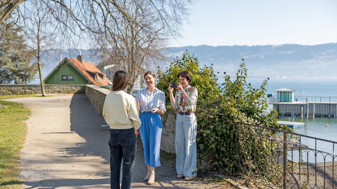 Drei Frauen unterhalten sich an einem Uferweg mit Blick auf einen See und Bergen im Hintergrund