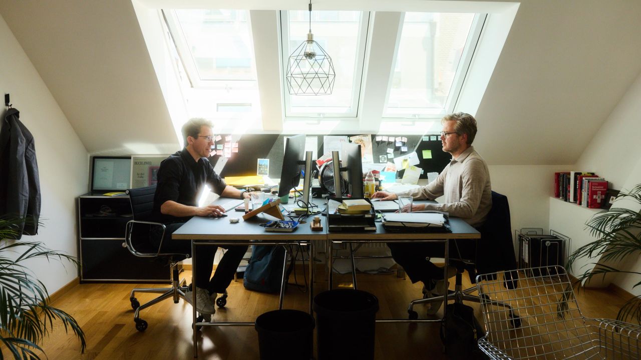 Zwei Männer sitzen sich an einem Schreibtisch mit mehreren Bildschirmen in einem Büro gegenüber