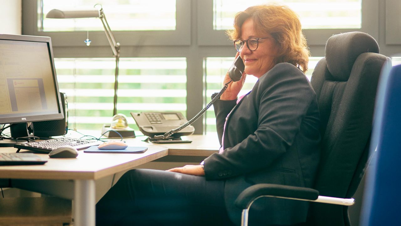 Frau in einem dunklen Business-Anzug telefoniert lächelnd am Schreibtisch in ihrem Büro