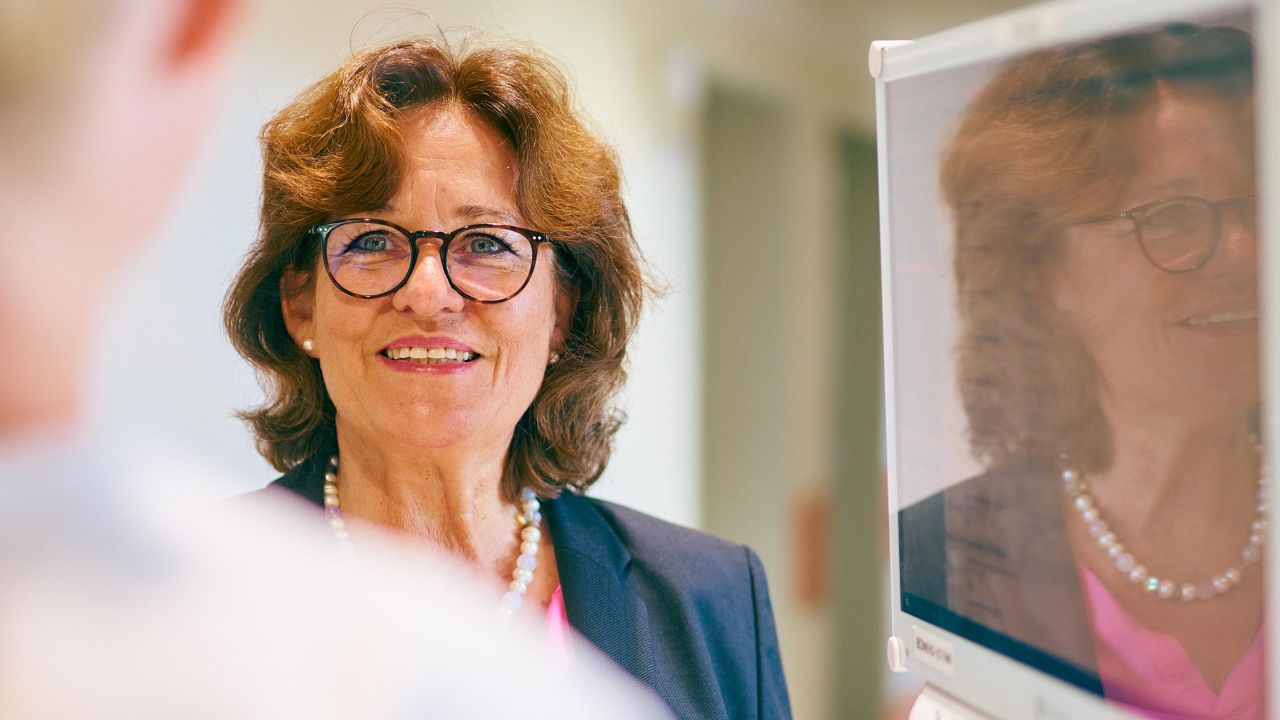 Lächelnde Frau mit Brille und Perlenkette im Gespräch, ihre Reflexion ist auf einem Monitor im Vordergrund sichtbar