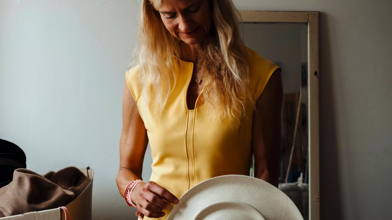 Eine blonde Frau in einem gelben Kleid hält einen hellen Hut mit beiden Händen vor dem Körper.