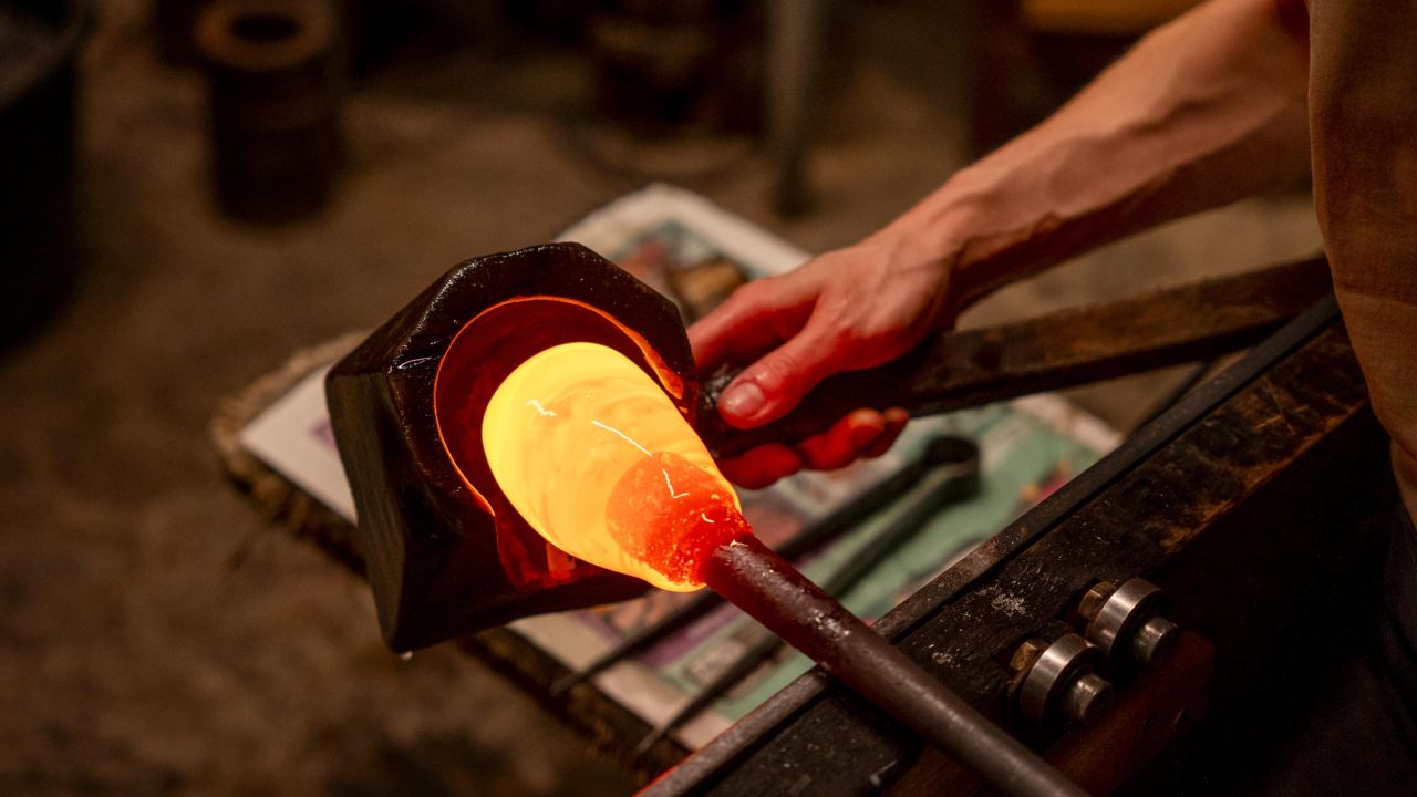 Detailaufnahme der Hand eines Glasbläsers, die mit einer Zange glühend heißes, orangefarbenes Glas an einer Glasmacherpfeife formt Detailaufnahme der Hand eines Glasbläsers, die mit einer Zange glühend heißes, orangefarbenes Glas an einer Glasmacherpfeife formt