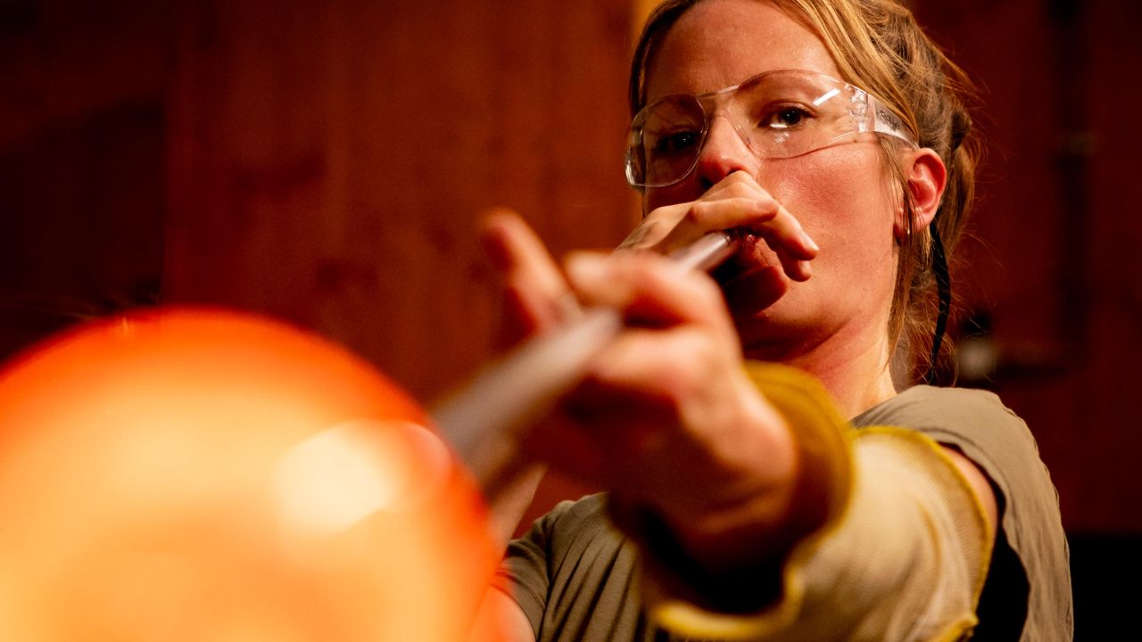Eine Glasbläserin mit Schutzbrille bläst durch eine lange Glasmacherpfeife, deren Spitze glühend orange leuchtet und unscharf im Vordergrund liegt Eine Glasbläserin mit Schutzbrille bläst durch eine lange Glasmacherpfeife, deren Spitze glühend orange leuchtet und unscharf im Vordergrund liegt