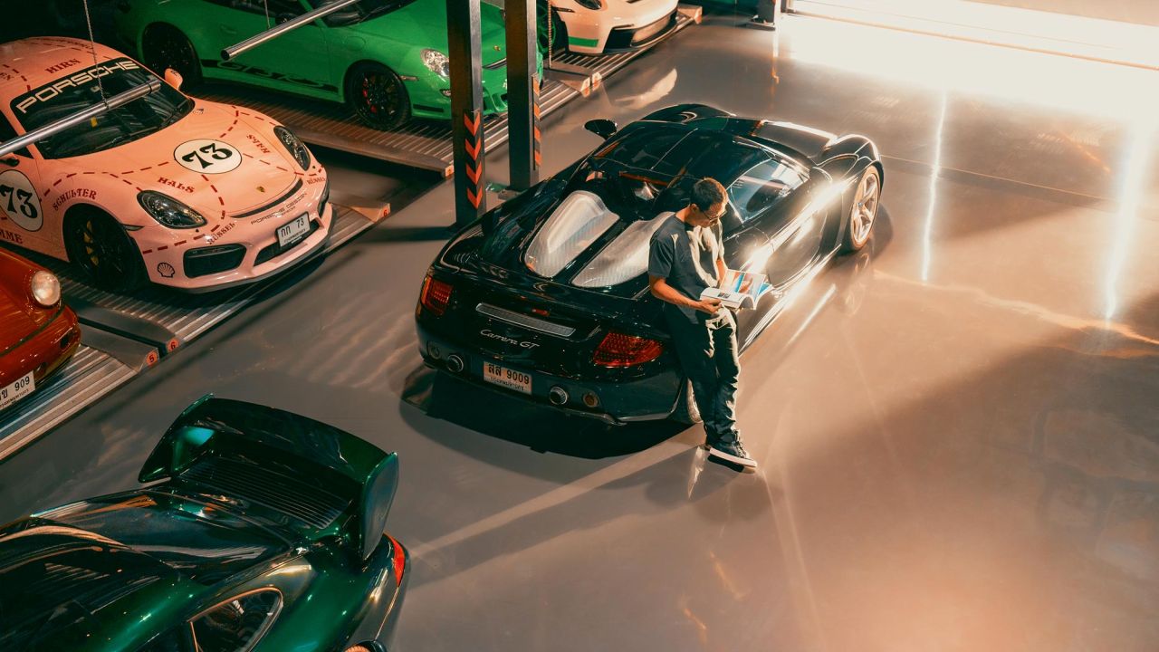 Mann lehnt in einer Garage an einem Carrera GT und blättert in Zeitschrift, umgeben von mehreren geparkten Porsche-Sportwagen