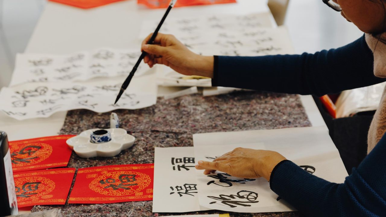 Frau sitzt an einem Tisch und schreibt chinesische Schriftzeichen mit einem Pinsel auf ein weißes Blatt Papier
