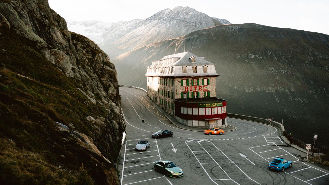 Historisches Hotel an einer scharfen Kurve einer Bergstraße im Alpenpanorama, auf dessen Parkplatz mehrere klassische Porsche-Fahrzeuge stehen Historisches Hotel an einer scharfen Kurve einer Bergstraße im Alpenpanorama, auf dessen Parkplatz mehrere klassische Porsche-Fahrzeuge stehen