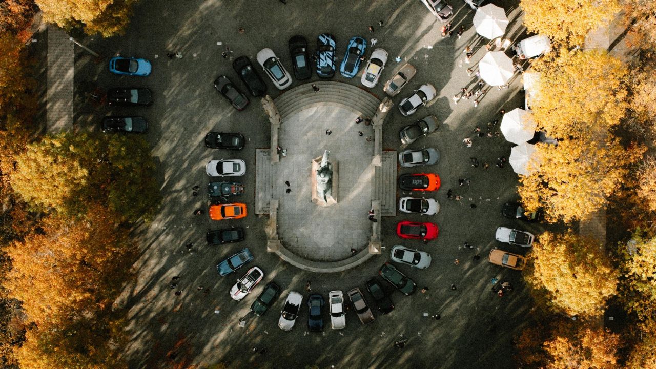 Drohnenaufnahme zeigt mehrere Porsche-Fahrzeuge, die im Kreis um ein Denkmal in einem von Bäumen gesäumten Parkplatz stehen Drohnenaufnahme zeigt mehrere Porsche-Fahrzeuge, die im Kreis um ein Denkmal in einem von Bäumen gesäumten Parkplatz stehen