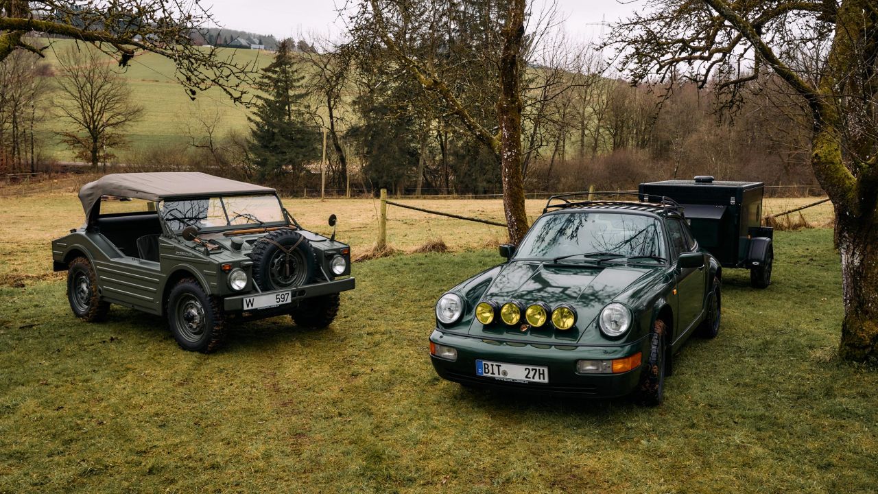 Ein dunkelgrüner Porsche 911 mit Zusatzscheinwerfern und Anhänger steht neben einem olivgrünen Militär-Geländewagen auf einer Wiese Ein dunkelgrüner Porsche 911 mit Zusatzscheinwerfern und Anhänger steht neben einem olivgrünen Militär-Geländewagen auf einer Wiese